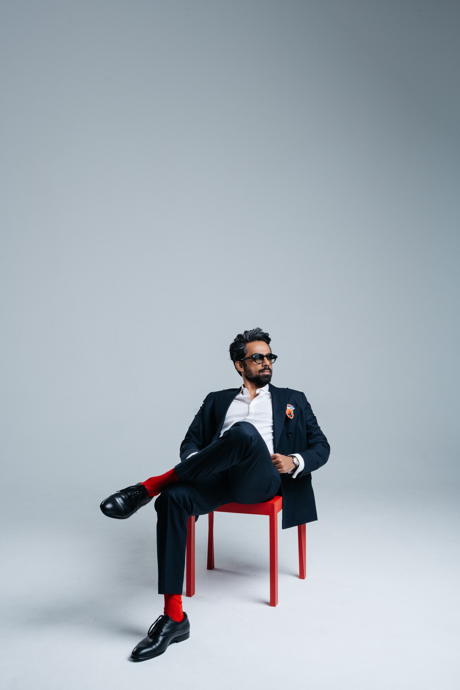 Kartik in a navy suit on a red chair, wearing red socks